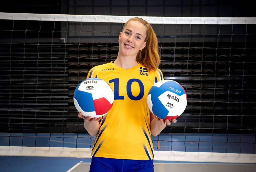 En tjej som står i en volleybollhall med en plan och nät i bakgrunden och en volleyboll i varje hand.